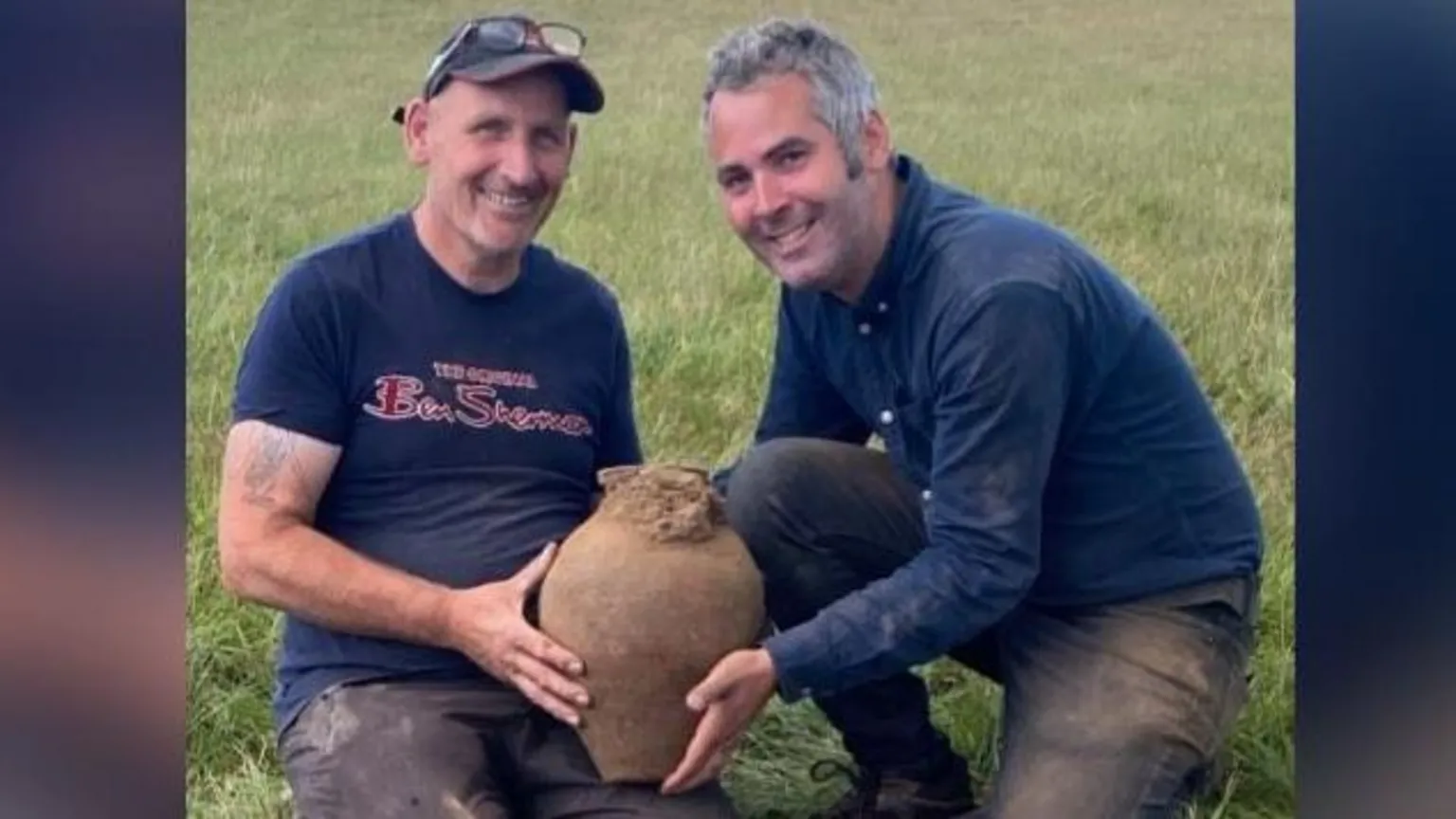 Minelab Two men holding a clay pot smiling with the dig site seen just in front of them. The man on the left has glasses on his head and a cap, he has a blue t-shirt with the words Ben Sherman, in red font, written on the front. The man on the right has salt and pepper hair, a blue shirt and blue pants. 